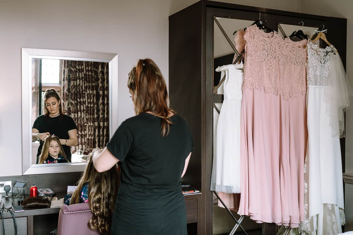 Flower girl having her hair done before the wedding ceremony with the bridesmaid's and bride's dresses hanging in the background